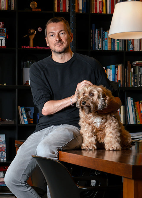 Floris van Bommel in a black long-sleeve shirt and light grey jeans sits on a wooden table, holding a small fluffy brown dog Flapper. He is positioned in front of a dark bookshelf filled with books, with a lamp visible on the right.