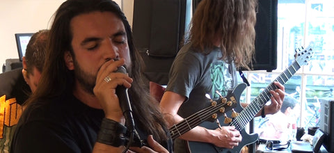 A live band performance featuring a vocalist with long dark hair singing into a microphone in the foreground, and a guitarist with long hair playing a black electric guitar in the background.