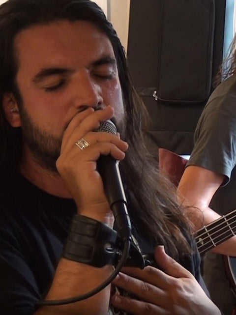 A close-up shot of a man with long dark hair and a beard singing into a microphone, wearing a black shirt and a black wrist cuff. Another person holding a bass guitar is partially visible in the background.