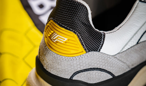 Visma sneaker Floris van Bommel. Close-up of the heel area of a low-top sneaker showing a yellow accent tab with a white stylised logo, alongside black mesh, gray suede, and white leather panels.