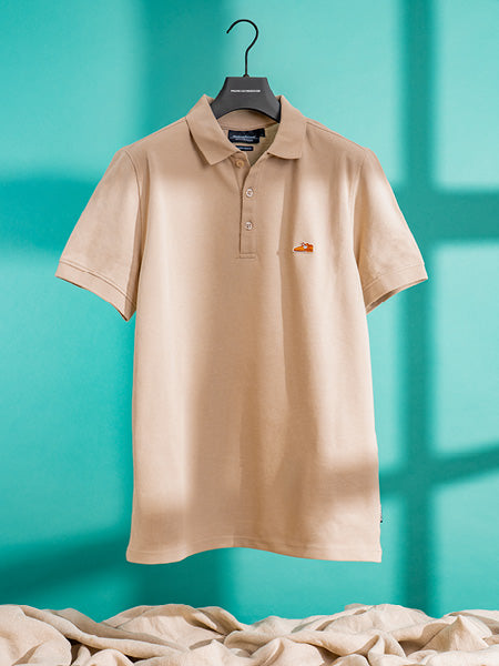 Floris van Bommel shirt. A beige short-sleeve polo shirt hanging on a black hanger against a turquoise background with soft window-shadow effects and draped fabric at the bottom.