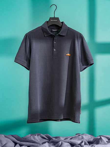 Floris van Bommel shirt. A dark charcoal gray polo shirt with a small embroidered logo on the left chest, hanging on a black hanger against a teal background with draped gray fabric at the bottom.