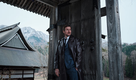 Floris van Bommel in a dark leather coat and blue suit stands in front of a large weathered wooden gate, with a traditional Japanese-style building and mountains in the background.
