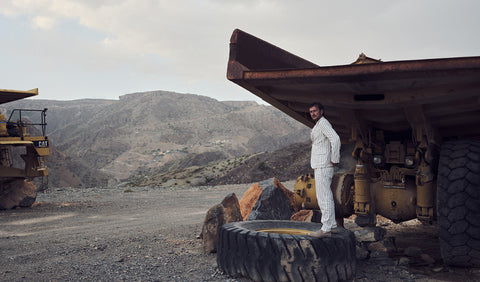 Floris van Bommel in a white suit stands on a large tyre at an open-pit mine, with a massive haul truck and rocky hills in the background.