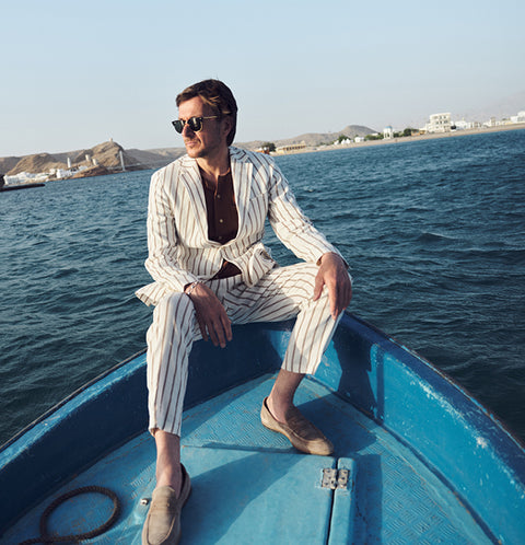 Floris van Bommel wearing a white pinstripe suit and sunglasses, sits in the blue prow of a boat on the water, with a coastal landscape in the background.