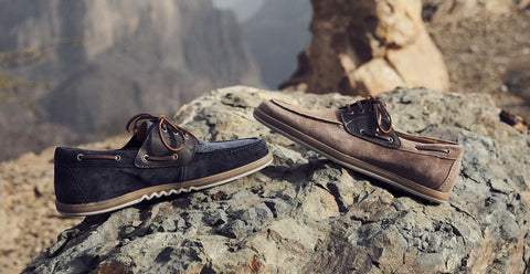 Two boat-style Floris van Bommel shoes resting on a large rock outdoors, with a blurred mountainous canyon landscape in the background. One shoe is dark navy suede with tan trim, and the other is taupe-brown suede with dark trim.