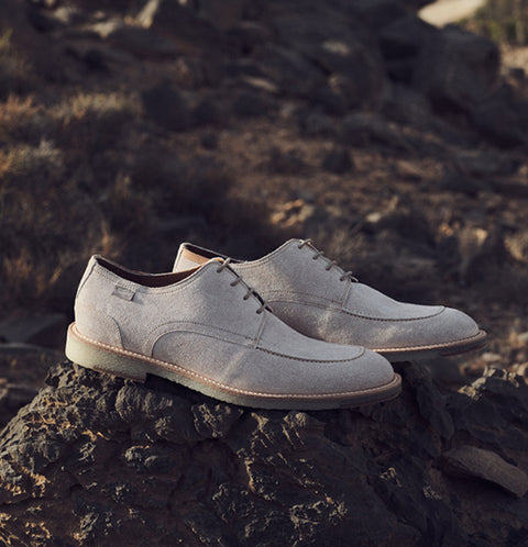 A pair of light grey suede Floris van Bommel shoes resting on a dark volcanic rock with a rocky, dry outdoor landscape in the background.