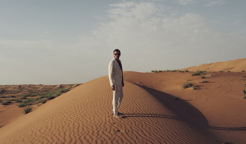 Floris van Bommel, wearing a light-colored pinstripe suit and sunglasses stands on a large sand dune in a desert landscape under a pale sky.