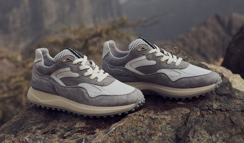 A pair of gray and off-white Floris van Bommel sneakers with textured panels and white laces, displayed on a large rock with a blurred mountainous outdoor background.