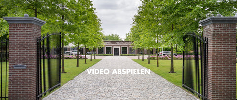 A wide outdoor view of a brick gated entrance with a paved driveway leading to a building, lined with green trees under a cloudy sky. Large white text across the center reads 'VIDEO ABSPIELEN'.