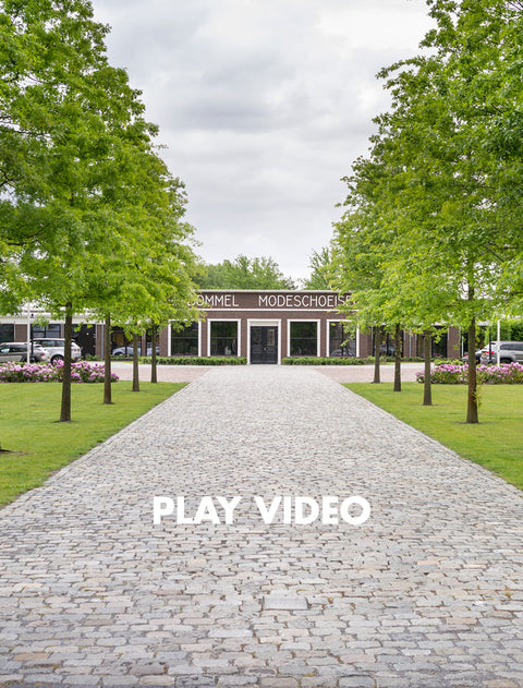 A paved pathway leads toward a building labeled 'Bommel' in a landscaped park setting with green trees and grass under a cloudy sky. Large white text reading 'PLAY VIDEO' is overlaid near the bottom centre.