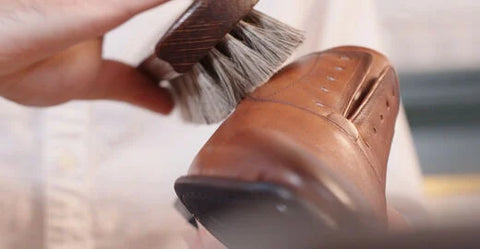 A close-up shot of a hand holding a brush and polishing a brown leather dress shoe. Floris van Bommel