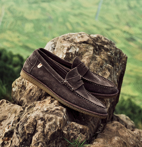 A pair of dark brown suede Floris van Bommel loafers resting on large rocks with a blurred green mountain landscape in the background.