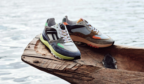 A pair of Floris van Bommel sneakers displayed on a rustic wooden log with water in the background. The shoes feature a mix of grey, navy, white, black, lime green, and orange accents.